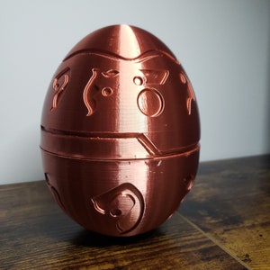 May include: A copper-colored egg-shaped object with intricate designs and a smooth, shiny finish. The egg is divided into two halves, with a seam running around the middle.