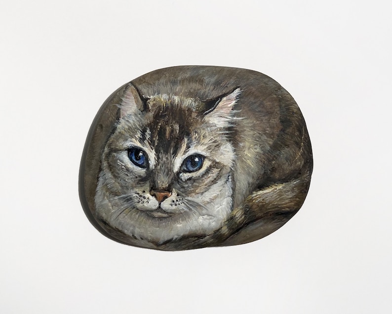 Puede incluir: Una piedra pintada a mano que representa un gato con ojos azules y pelaje gris y marr&oacute;n. El gato est&aacute; acurrucado, con la cabeza y la cara en el centro de la piedra. La piedra tiene forma ovalada y est&aacute; sobre un fondo blanco.