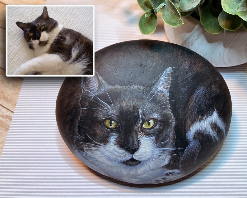 Puede incluir: Una piedra pintada a mano, de forma ovalada, presenta un gato blanco y negro con ojos amarillos. El retrato del gato est&aacute; detallado con pelaje y rasgos faciales realistas. Una foto m&aacute;s peque&ntilde;a del gato est&aacute; en la esquina superior izquierda. La piedra est&aacute; sobre una superficie rayada.