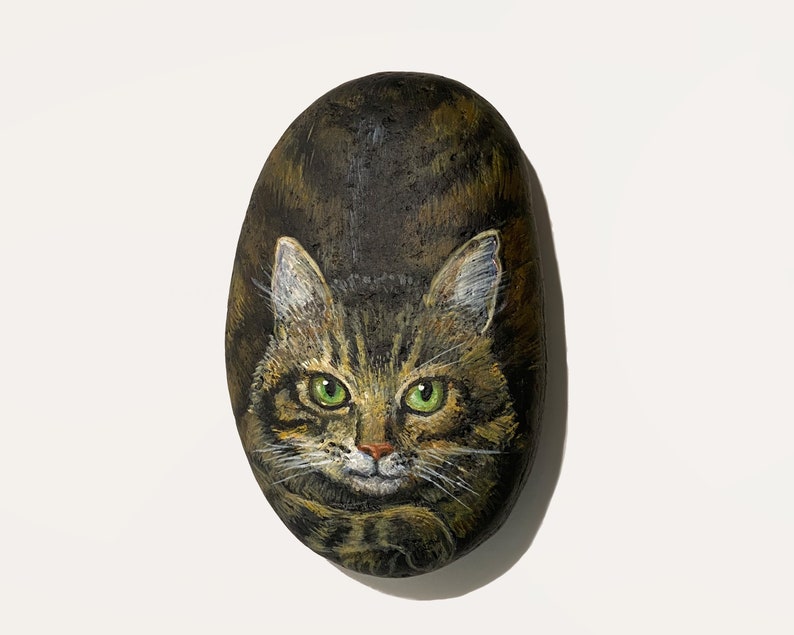 Puede incluir: Piedra pintada a mano con un gato atigrado de ojos verdes. La cara del gato est&aacute; centrada en una piedra ovalada de color marr&oacute;n oscuro. La obra de arte presenta pinceladas detalladas y patrones de pelaje realistas.