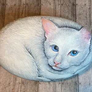 Puede incluir: Piedra pintada a mano que representa un gato blanco con ojos azules y orejas y nariz rosas. El gato est&aacute; acurrucado sobre una piedra de forma ovalada. La obra de arte es detallada y realista, con un aspecto suave y texturizado.