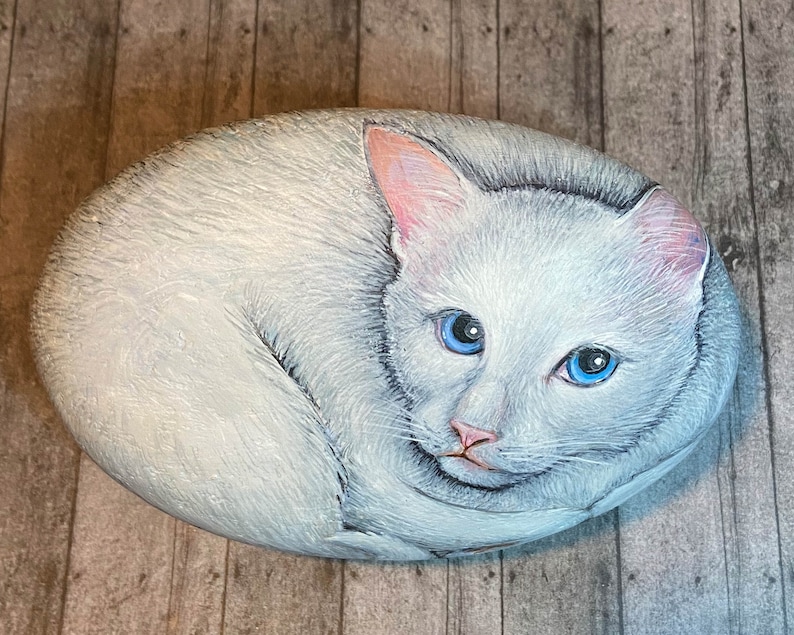 Puede incluir: Piedra pintada a mano que representa un gato blanco con ojos azules y orejas y nariz rosas. El gato est&aacute; acurrucado sobre una piedra de forma ovalada. La obra de arte es detallada y realista, con un aspecto suave y texturizado.