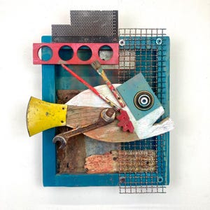 May include: A colorful, mixed-media fish sculpture made from wood, metal, and paint. The fish has a yellow tail, a blue eye, and a red mouth. The sculpture is framed in a teal blue frame with a wire mesh background.