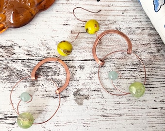 May include: Copper wire earrings with green and yellow glass beads. The earrings are handmade and feature a unique design.