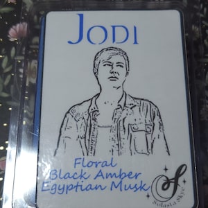 May include: A white rectangular product label with the name "JODI" in blue script above a black and white illustration of a person. Below the illustration are the words "Floral Black Amber Egyptian Musk". The product is from Solasta Skye.
