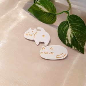 May include: Two white ceramic cat pins with gold outlines. The cats are in different poses, one sitting and one lying down.