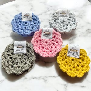 May include: Set of four crocheted coasters in blue, grey, pink, and yellow. Each coaster has a circular design with a scalloped edge. The coasters are handmade and have a tag that says "Coasters - 100% Handmade".