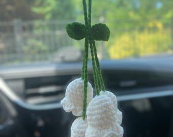 May include: A handmade crochet car charm featuring three white lily of the valley flowers. The flowers are suspended from green stems and a green bow. The background shows a blurred view of trees and a blue sky.