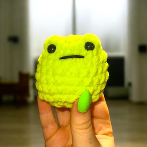 May include: A bright yellow crocheted frog toy with black eyes. The frog is being held by a hand with green nail polish.
