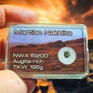 Könnte beinhalten: Ein kleiner, durchsichtiger Kunststoffbehälter mit einer Probe des Marsmeteoriten Nakhilite. Der Behälter trägt die Aufschrift "Martian Nakhilite", "NWA 15200", "Augite-rich" und "TKW 196g". Die Probe ist ein kleiner, grün gefärbter Stein.