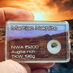 Könnte beinhalten: Eine transparente Kunststoffhülle mit einem Exemplar von Martian Nakhilite. Auf der Hülle steht "Martian Nakhilite", "NWA 15200", "Augite-rich" und "TKW 196g". Das Exemplar ist ein kleiner, grün-grauer Stein, der 196 Gramm wiegt.