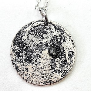 Op de afbeelding: Zilverkleurige ketting met een gedetailleerde maan-hanger. De hanger toont het oppervlak van de maan met kraters en een kleine, donkere steen. De ketting is zilver. Een uniek sieraad, geschikt voor dagelijks gebruik.
