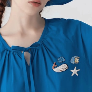 以下が含まれることがあります： マリンをテーマにした4つのブローチが付いた青いトップス。ブローチには、赤いアクセントが付いた白いクジラ、青い魚、白いヒトデ、青い貝殻が含まれています。トップスはネックラインにリボンが付いています。