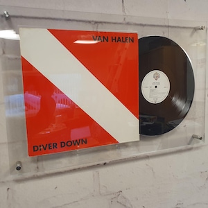 May include: A Van Halen Diver Down vinyl record and album cover displayed in a clear acrylic wall mount. The square cover features a red background with a white diagonal stripe and black text. The black vinyl record is positioned to the right.