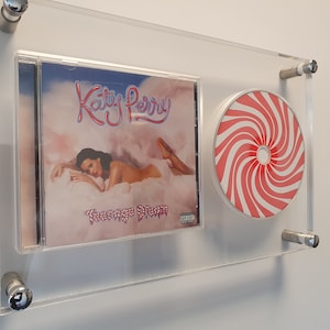 Puede incluir: Un CD enmarcado con una portada rosa y azul con el texto "Katy Perry" y el título "Teenage Dream". También se enmarca un disco rojo y blanco con forma de bastón de caramelo.