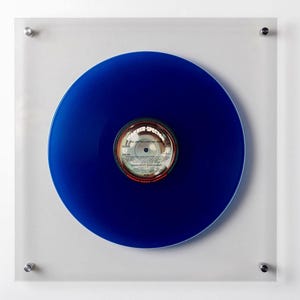 May include: A vibrant blue vinyl record displayed within a clear acrylic frame. The record's label is visible at the center, featuring text and a circular design. The frame is mounted on a white wall, secured by silver hardware.