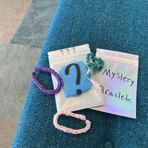 May include: A collection of colorful woven bracelets, including purple, pink, and a green and blue combination, are displayed. Two clear bags are labeled with "Mystery Bracelets" and "???" with a blue item and a question mark.