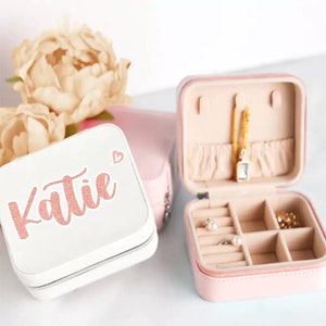 May include: A white and pink jewellery box with a zipper closure. The top of the box has the name "Katie" written in pink glitter. The bottom of the box has compartments for storing jewellery.