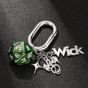 以下が含まれることがあります： 緑色の20面ダイス、ドラゴンチャーム、星型チャーム、そして「Wick」と書かれたシルバーチャームが付いたシルバーキーチェーンです。