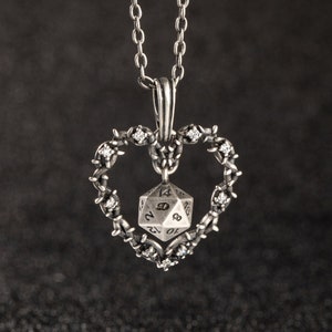 Puede incluir: Un collar de plata con un colgante en forma de corazón que presenta un dado de 20 caras de plata y cristales transparentes. El corazón está adornado con un diseño de alambre de púas.
