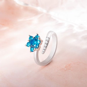 Könnte beinhalten: Ein silberner Ring mit einem blauen Edelstein in der Mitte, umgeben von kleineren blauen Edelsteinen. Der Ring ist offen und hat eine einzelne Reihe kleiner, klarer Edelsteine auf der Band.