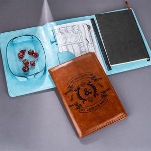 Könnte beinhalten: Ein braunes Ledernotizbuch mit einem blauen und weißen Umschlag. Das Notizbuch hat ein Würfelsymbol und den Text "The Dice Giveth On Jason's Matt".