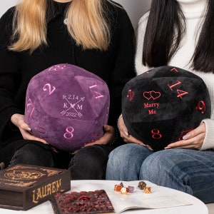May include: Two plush dice with embroidered designs. The purple dice has the date 2.14.2022, the initials K and M, and an arrow pointing to a heart. The black dice has two hearts and the text "Marry Me".