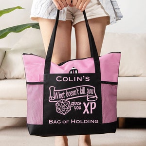 Könnte beinhalten: Eine rosa und schwarze Tragetasche mit dem Text "COLIN'S What doesn't kill you gives you XP BAG OF HOLDING" und einem rosa und weißen 20-seitigen Würfel.