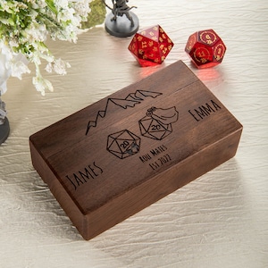 May include: A wooden box with a mountain and dice design. The box is engraved with the names "James" and "Emma" and the text "Roll Mates Est 2022".