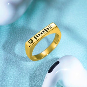Puede incluir: Un anillo de color dorado con una banda cuadrada. La parte superior de la banda tiene un diseño de código de barras en blanco y negro con un pequeño símbolo circular al principio.