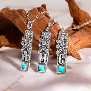 May include: Three silver necklaces with turquoise stones and floral designs. Each pendant features a different letter: A, M, and C.