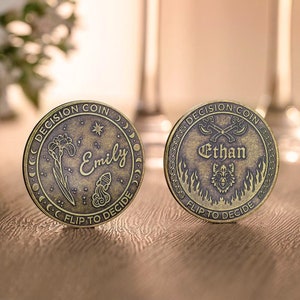 May include: Two antique gold-toned decision coins with the names "Emily" and "Ethan" engraved on them. The Emily coin features a flower and a star, while the Ethan coin features a wolf and crossed axes. Both coins have the text "Decision Coin" and "Flip to Decide" around the edges.