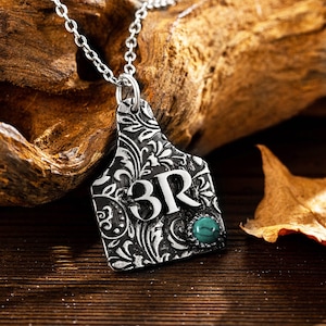 May include: Silver necklace with a silver pendant shaped like a cattle ear tag. The tag has an ornate floral design and the initials "3R" in the center. A turquoise stone is set in the bottom of the tag.