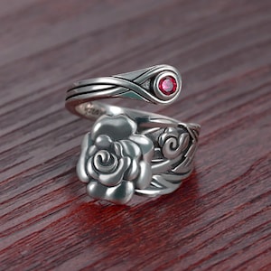 May include: Silver ring with a rose design and a red gemstone. The ring is open and wraps around the finger.