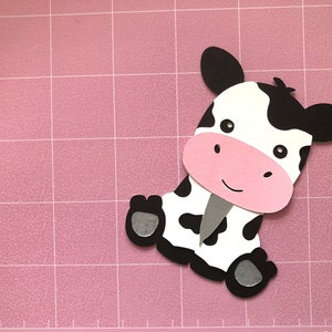 Puede incluir: Un recorte de papel en blanco y negro de una vaca con la nariz rosa y los ojos negros. La vaca est&aacute; sentada en una estera de corte rosa.