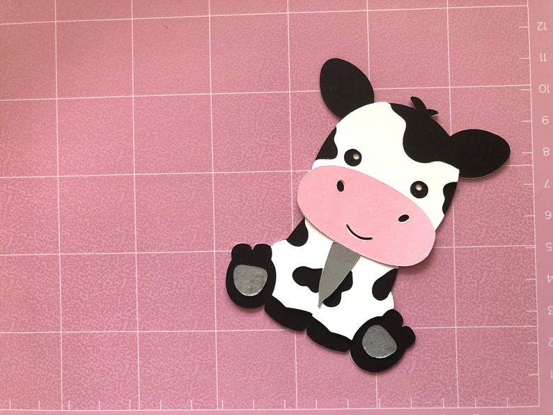 Puede incluir: Un recorte de papel en blanco y negro de una vaca con la nariz rosa y los ojos negros. La vaca est&aacute; sentada en una estera de corte rosa.