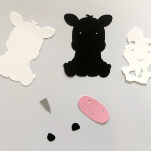 Puede incluir: Un conjunto de recortes de papel para un proyecto de manualidades. El conjunto incluye dos recortes de vaca blancos, un recorte de vaca negro, una nariz rosa, dos ojos negros y un cuerno gris.