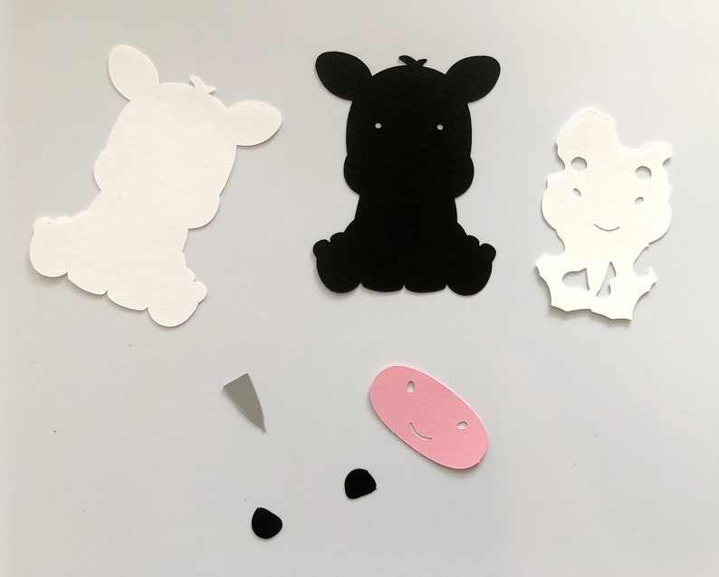 Puede incluir: Un conjunto de recortes de papel para un proyecto de manualidades. El conjunto incluye dos recortes de vaca blancos, un recorte de vaca negro, una nariz rosa, dos ojos negros y un cuerno gris.