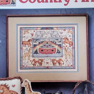 May include: A craft pattern book titled "Country Ark" by Susan Winget. The cover features a framed cross-stitch design of Noah's Ark with animals. Decorative cross-stitch animal figures are displayed on a wooden shelf.