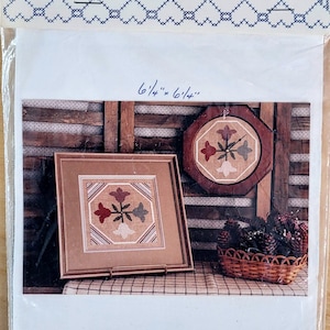 May include: A cross-stitch pattern featuring a floral design in shades of red, green, and white. The pattern is framed and displayed on a wooden wall with a basket of pine cones. The pattern is 6 1/4 inches by 6 1/4 inches.