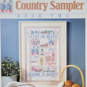 May include: A craft book titled "Country Sampler Book Two" by Nancy Rossi. The cover features a framed cross-stitch sampler with the text "East or West Home is Best". An embroidery hoop, scissors, thread, and a basket of bread are also visible.