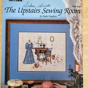 May include: A vintage sewing pattern book titled "The Upstairs Sewing Room" by Paula Vaughan. The cover features a framed cross-stitch design of a sewing room, with spools of thread, buttons, and a pincushion in the foreground. The book is a leaflet from Leisure Arts.