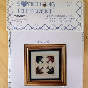 May include: A framed cross-stitch pattern with a geometric design in shades of black, red, and beige. The frame is a warm brown color. The package includes the text "SOMETHING DIFFERENT" and the dimensions 6" x 6 1/4".