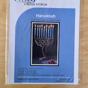 May include: A cross stitch pattern for a Hanukkah menorah with nine blue candles lit. The menorah is silver with a black background. The pattern is by Tereena Clarke and is called "Hanukkah".