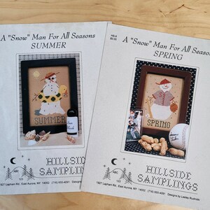 May include: Two cross-stitch patterns featuring a snowman in different seasonal settings. The first pattern shows a snowman in a yellow life preserver with a beach scene, titled "A "Snow" Man For All Seasons SUMMER". The second pattern shows a snowman in a baseball uniform with a baseball and peanuts, titled "A "Snow" Man For All Seasons SPRING". Both patterns are from Hillside Samplings.