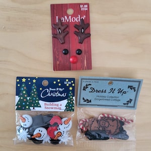 May include: A collection of Christmas-themed embellishments, including reindeer-shaped buttons, snowman-themed buttons, and gingerbread cottage buttons. The reindeer buttons are on a red and brown card. The snowman buttons are in a clear plastic bag.