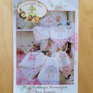May include: A brochure featuring a collection of Cottage Flowerpot Tea Towels. The image showcases several tea towels with floral embroidery, alongside plates and a teapot. The text "Crab-apple Hill" and "#232 Cottage Flowerpot Tea Towels" are visible.