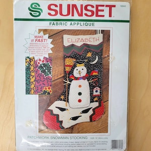 May include: A Sunset Fabric Applique kit for a patchwork snowman stocking. The kit includes embroidery thread, fabric, and instructions. The stocking is 16 inches long and features a snowman with a black hat, a red scarf, and a black bird design. The stocking is personalized with the name "Elizabeth".