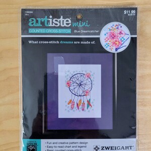 May include: A counted cross-stitch kit featuring a blue dreamcatcher with colorful feathers and flowers. The kit is called "artiste mini" and is designed by "Kooler Design Studio".
