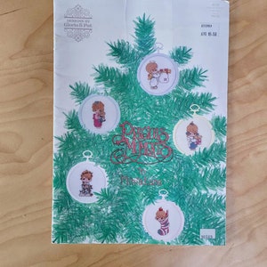 May include: A cross-stitch pattern booklet featuring Precious Moments ornaments. The cover shows a green Christmas tree with five ornaments, each depicting a different Precious Moments character. The title of the booklet is "Precious Moments in Miniature".